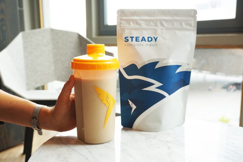 A hand holding a Queal shaker with an orange lid, filled with a Queal shake, next to a pouch of Queal Steady complete meals, placed on a marble table with a window in the background.