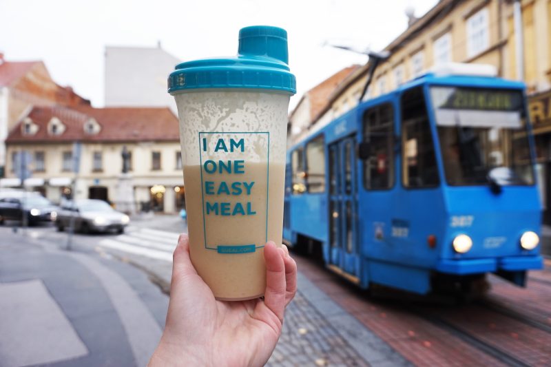 A hand holding a Queal shaker with a blue lid, filled with a Queal shake, with the text "I AM ONE EASY MEAL" on the shaker, in front of a city street scene with a blue tram and buildings.